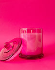 A beautiful hot pink glass cylinder shaped candle sits in the middle of the page. The glass lid with a round knob sits on the left hand side at an angle resting on the candle. You can see the white wick through the glass. The background of the image is also a beautiful hot pink color.