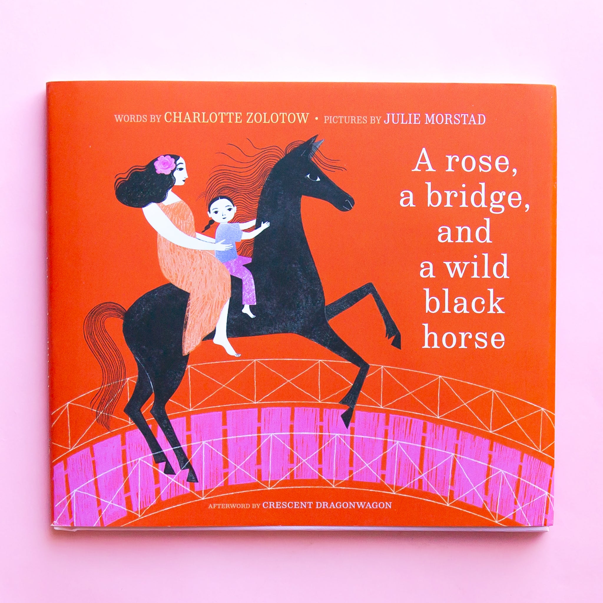 On a pink background is a red book with an illustration of a mother and child on a black horse and a white title that reads, &quot;A rose, a bridge, and a wild black horse&quot;. 
