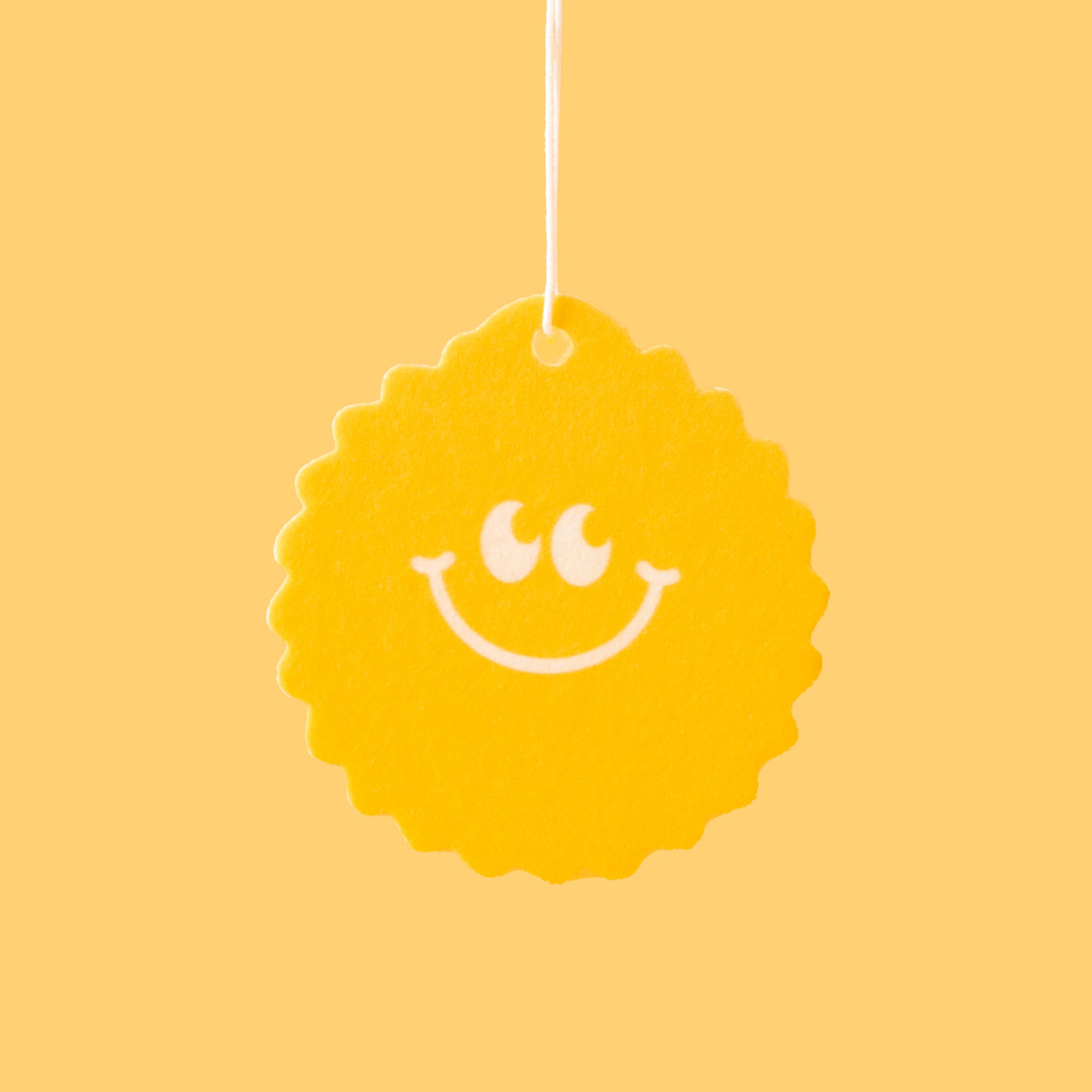 On a yellow background is a yellow sunshine shaped air freshener with a smiley face and a white hanging string. 