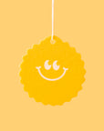 On a yellow background is a yellow sunshine shaped air freshener with a smiley face and a white hanging string.