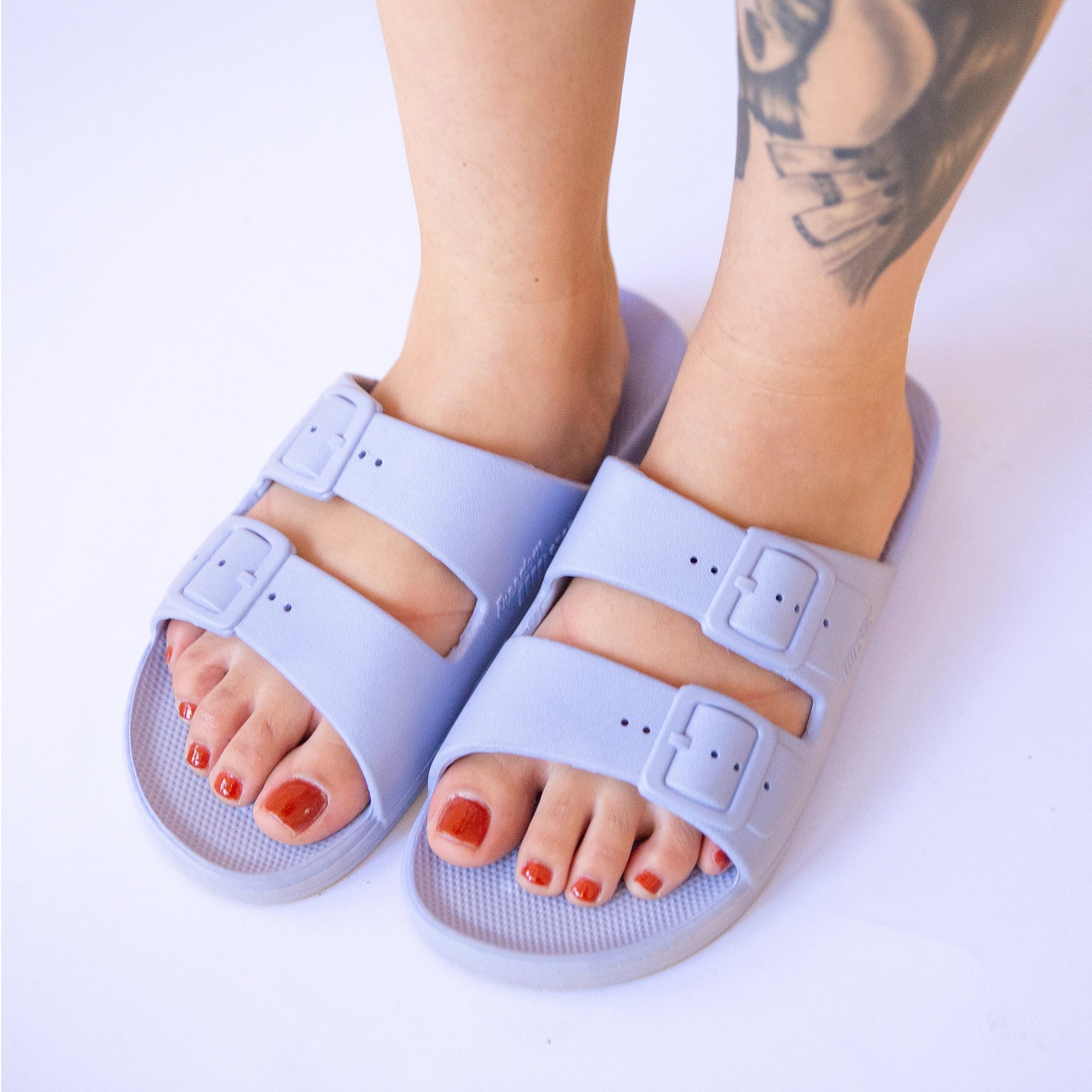 On a white background is a pair of light lavender cloud slides with two buckle straps on a model. 