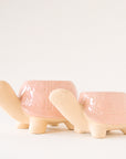 On a light grey background is two different sized ceramic planters in the shape of a turtle with a light pink "shell" with a subtle floral print.