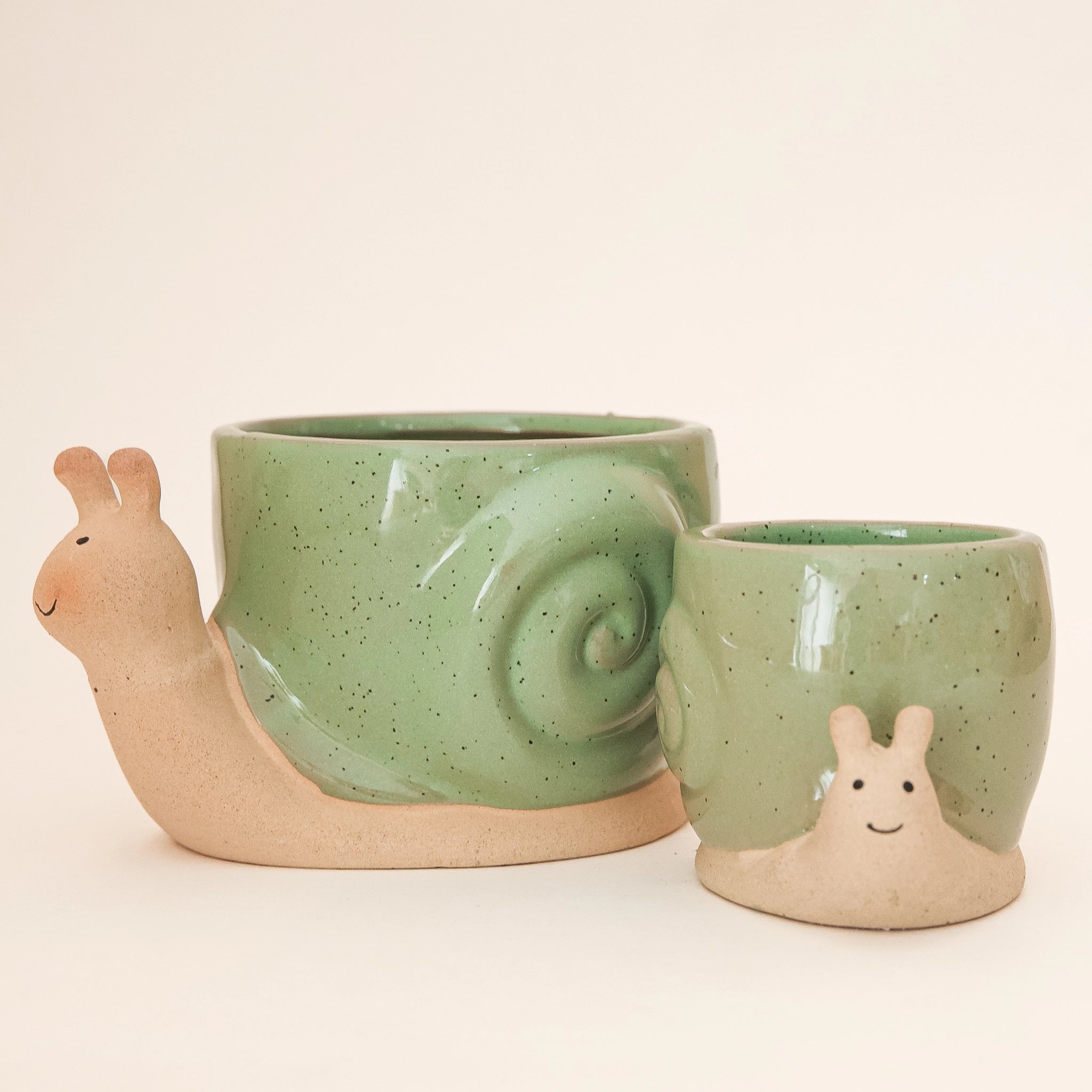 On a tan background is two ceramic planters in the shape of a snail with smiling faces and a swirly green &quot;shell&quot;.