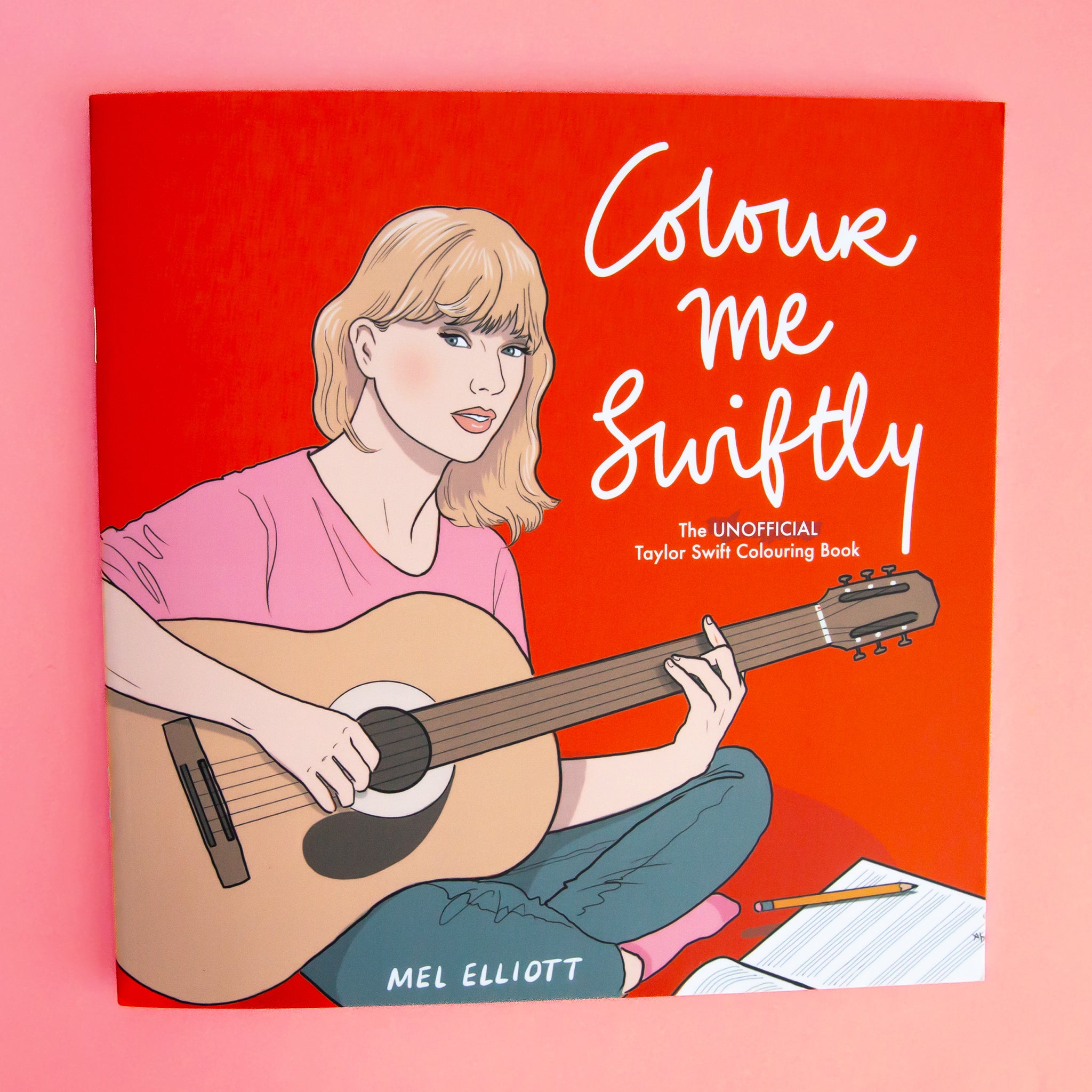 On a pink background is a red book with an illustration of Taylor Swift with a guitar and the white title on the right that reads, &quot;Colour me Swiftly The Unofficial Taylor Swift Colouring Book&quot;.