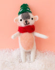 On a bright pink background is a white and pink felt ornament in the shape of a dog with a green hat and a red scarf.
