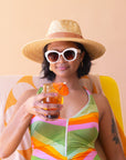 On a cream background is a model wearing a light honey colored straw sun hat with a tan ribbon around the base.