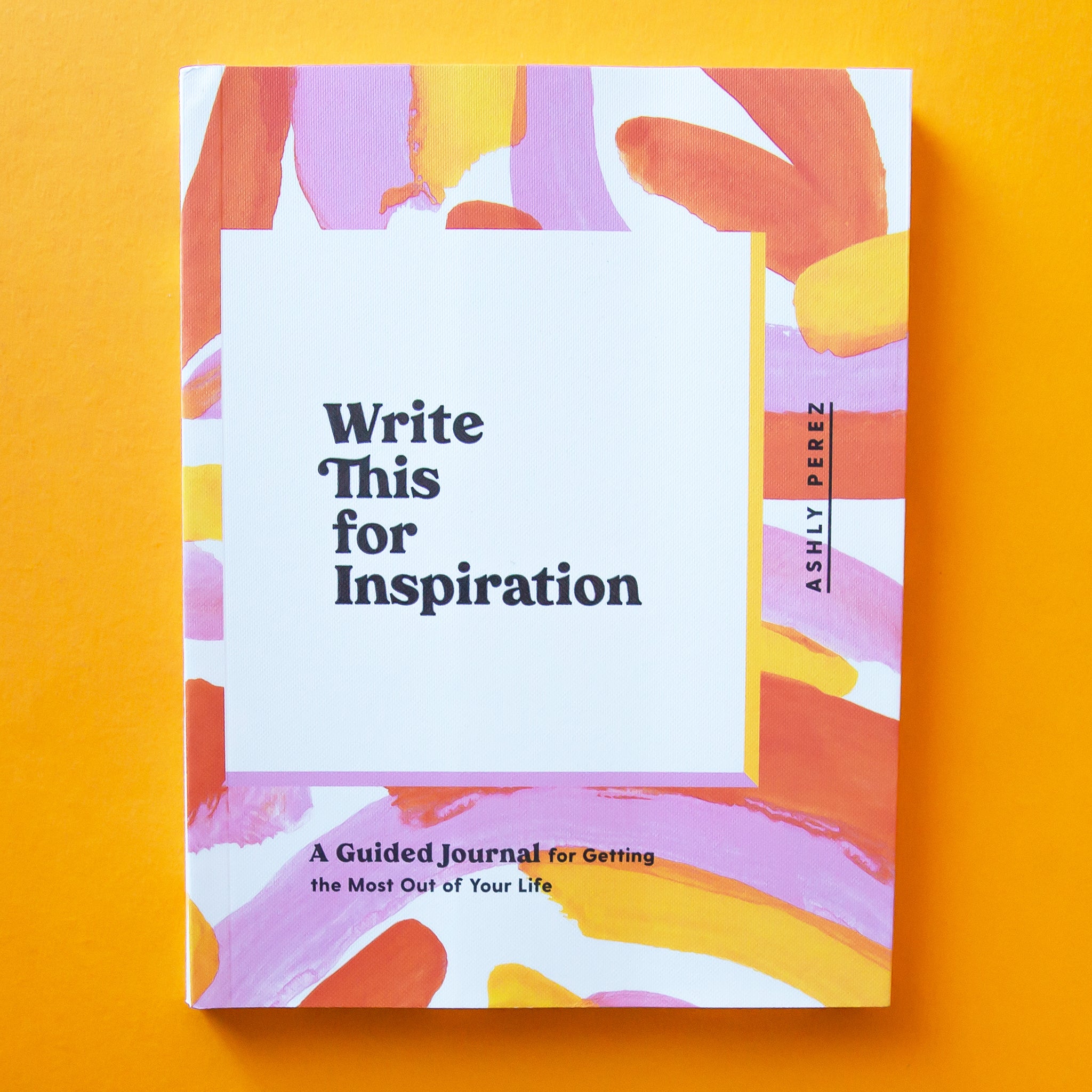 This journal is titled &#39;Write This for Inspiration by Ashly Perez&#39; in black lettering and has an enjoyable free-hand painted design with shades of pinks and oranges on top of a white background. The binding of the book is also white with the title spelt out in similar colorful lettering.