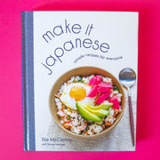 On a pink background is an ivory book cover with a book of food and wavy text above it that reads, "make it japanese simple recipes for everyone".