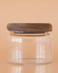 On a peach background is a clear glass storage canister with an acacia wood lid.