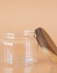 On a peach background is a clear glass storage canister with an acacia wood lid.