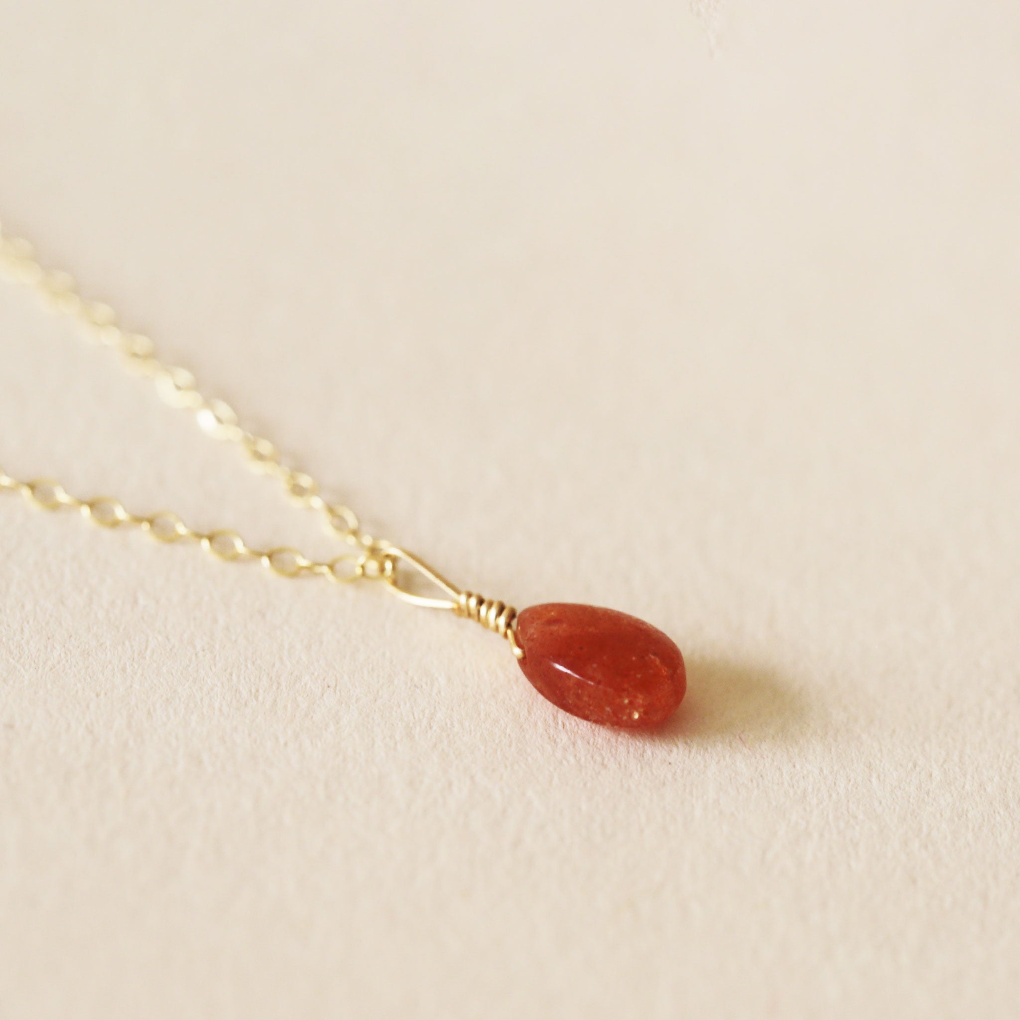 On a neutral background is a gold chain necklace with an orange/red Sunstone stone in the center.