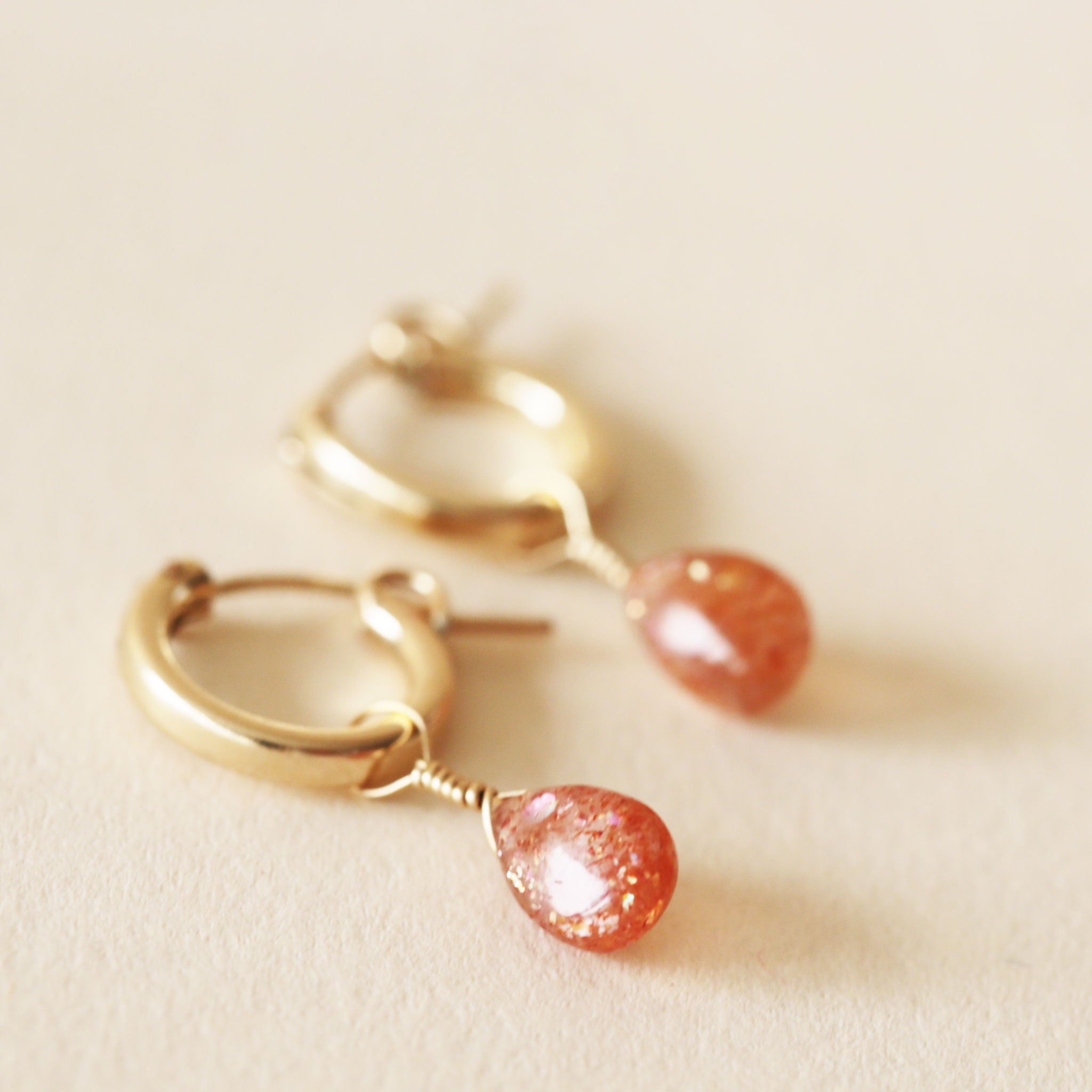 On a neutral background is a gold pair of huggie hoop earrings with a Sunstone drop gem detail.