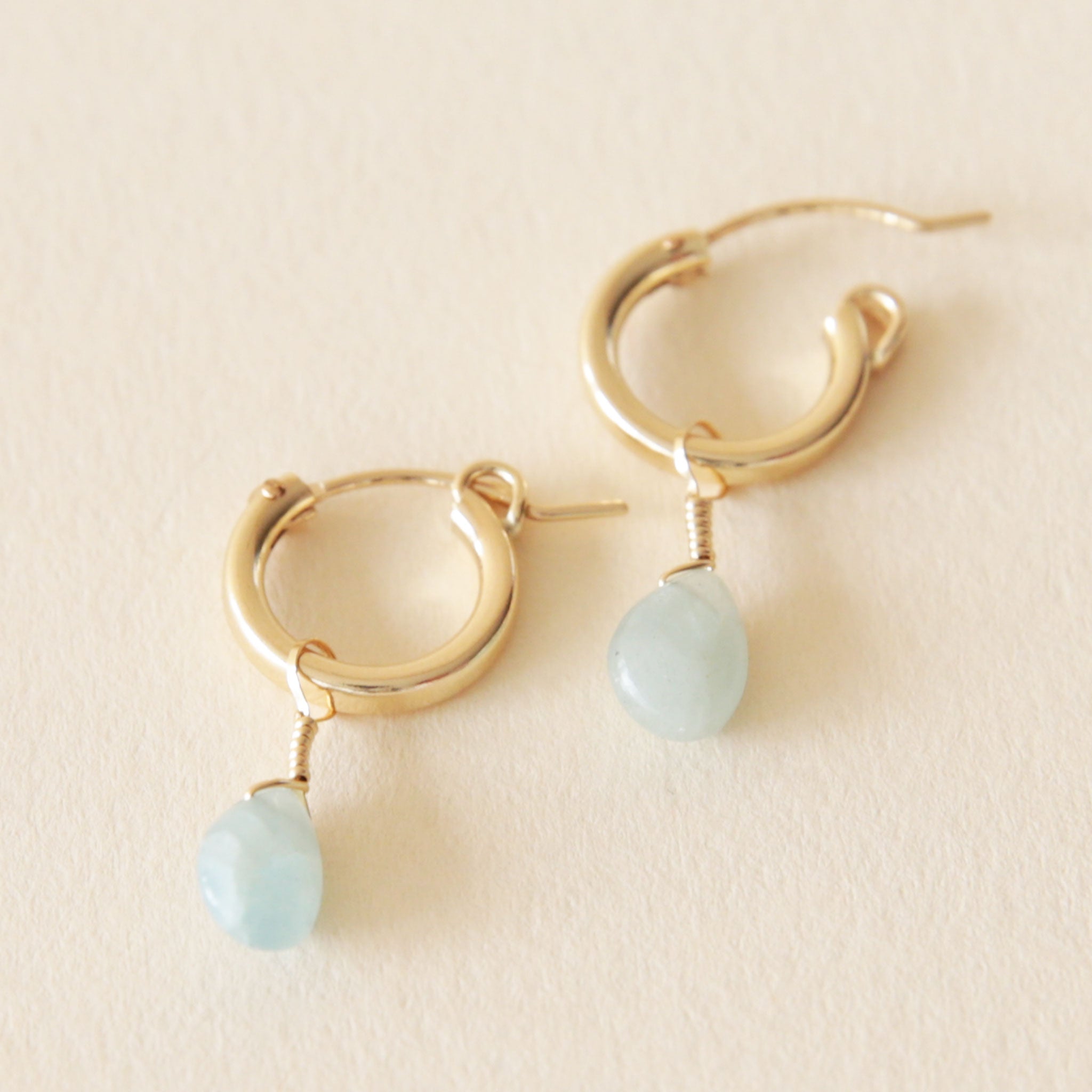 On a neutral background is a gold pair of huggie hoop earrings with a blue Aquamarine stone dangle deal.