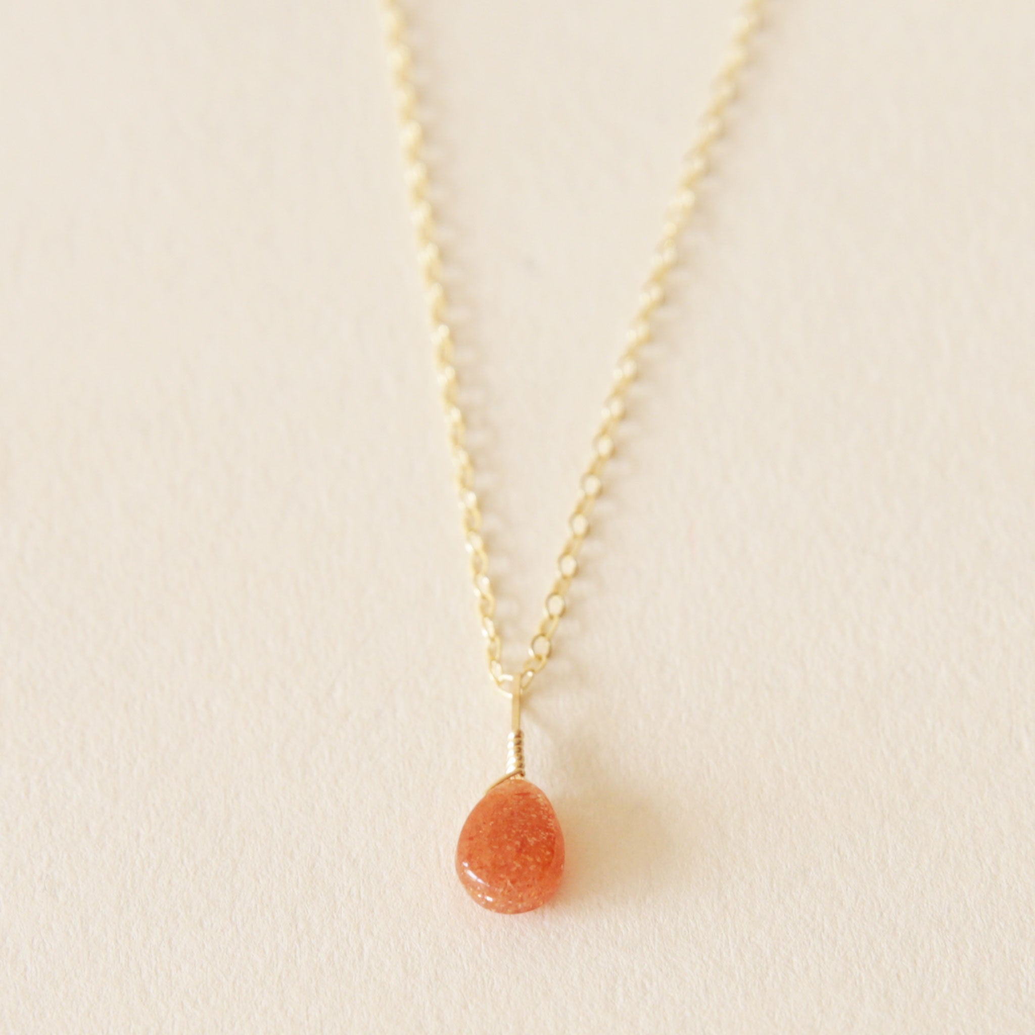 On a neutral background is a gold chain necklace with an orange/red Sunstone stone in the center. 