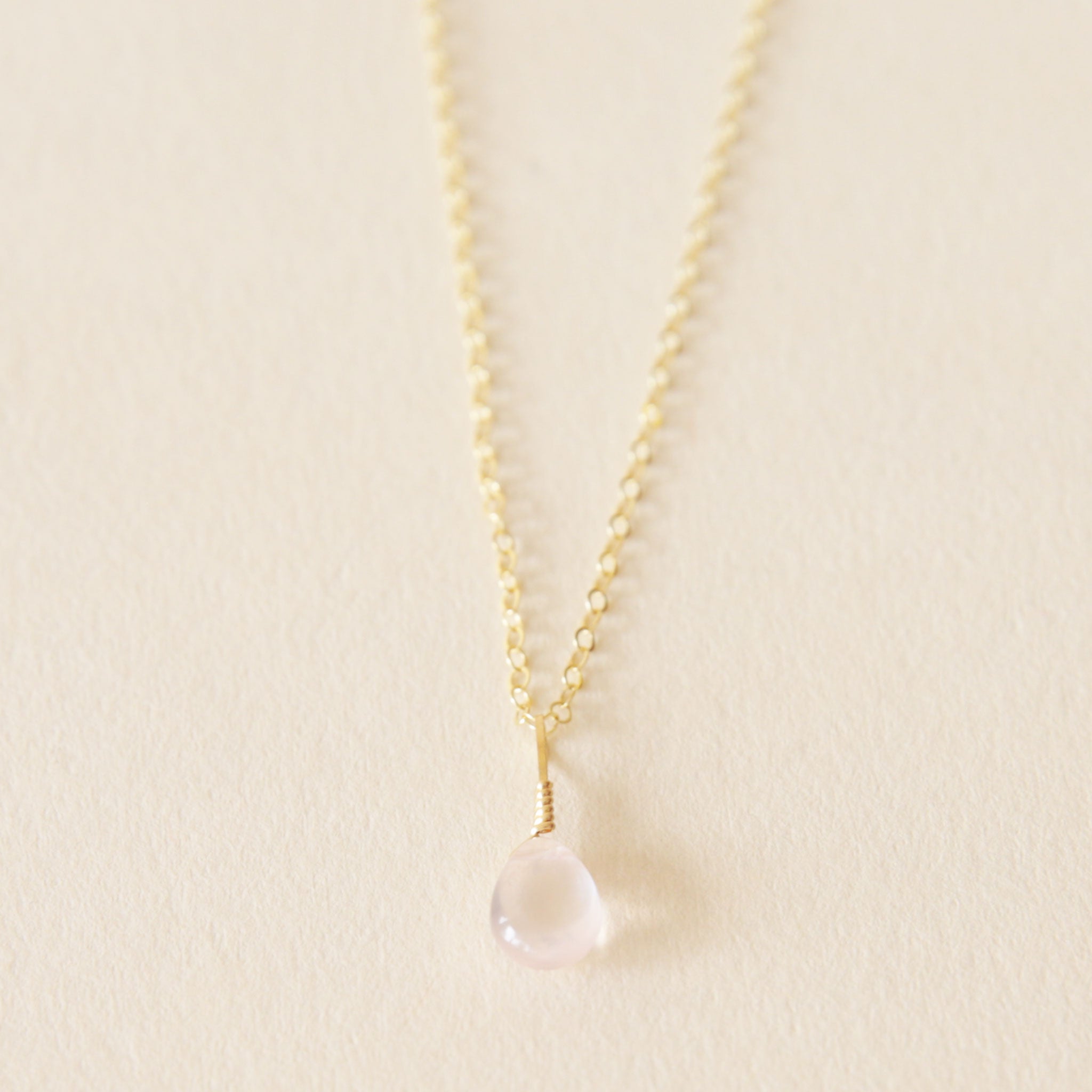 On a neutral background is a gold chain necklace with a rose quartz stone in the center.