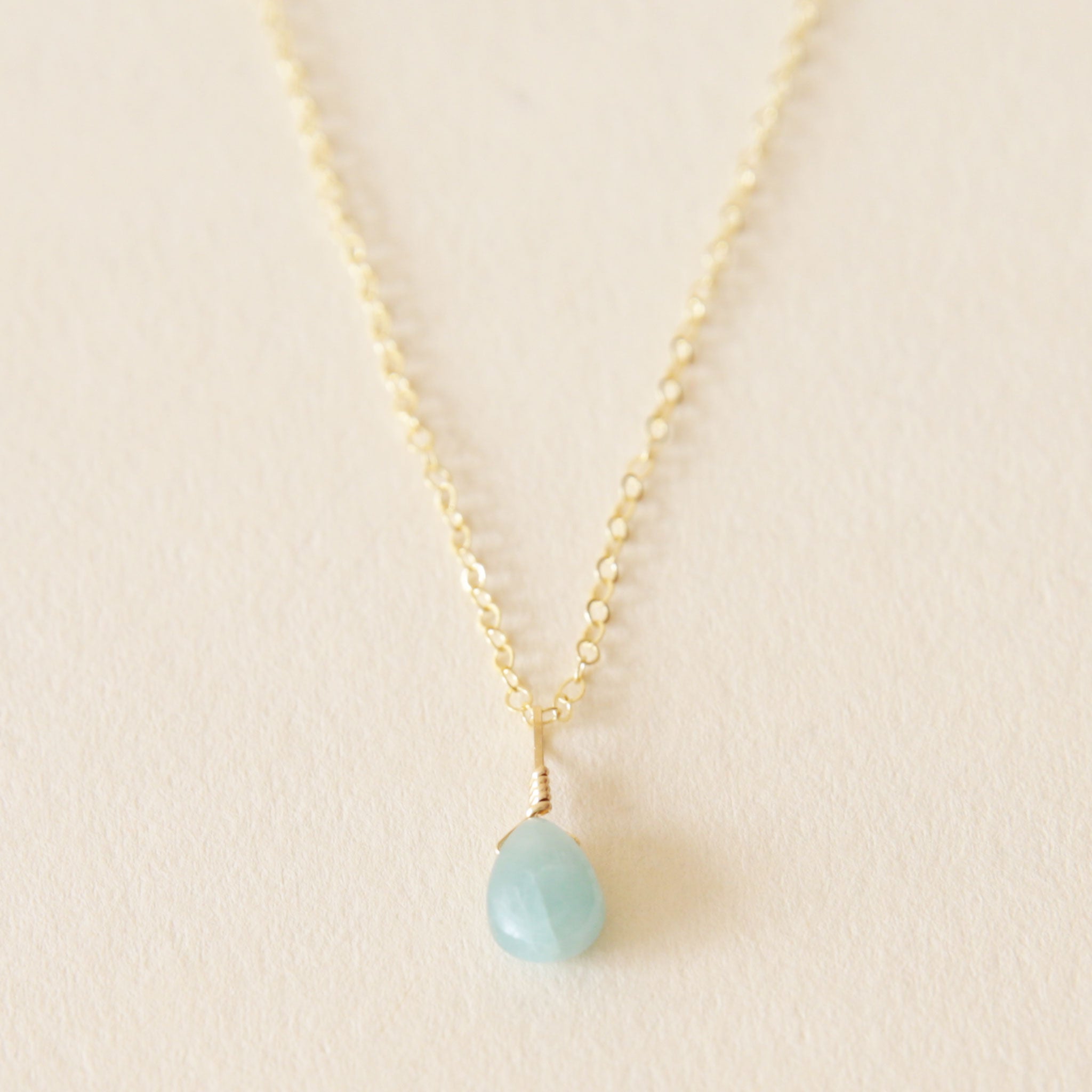 On a neutral background is a gold chain necklace with a blue Aquamarine stone at the center.