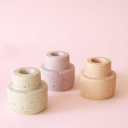 On a pink background is three ceramic speckled candle stick holders in three different shades. From left to right there is off white, pink and peach.