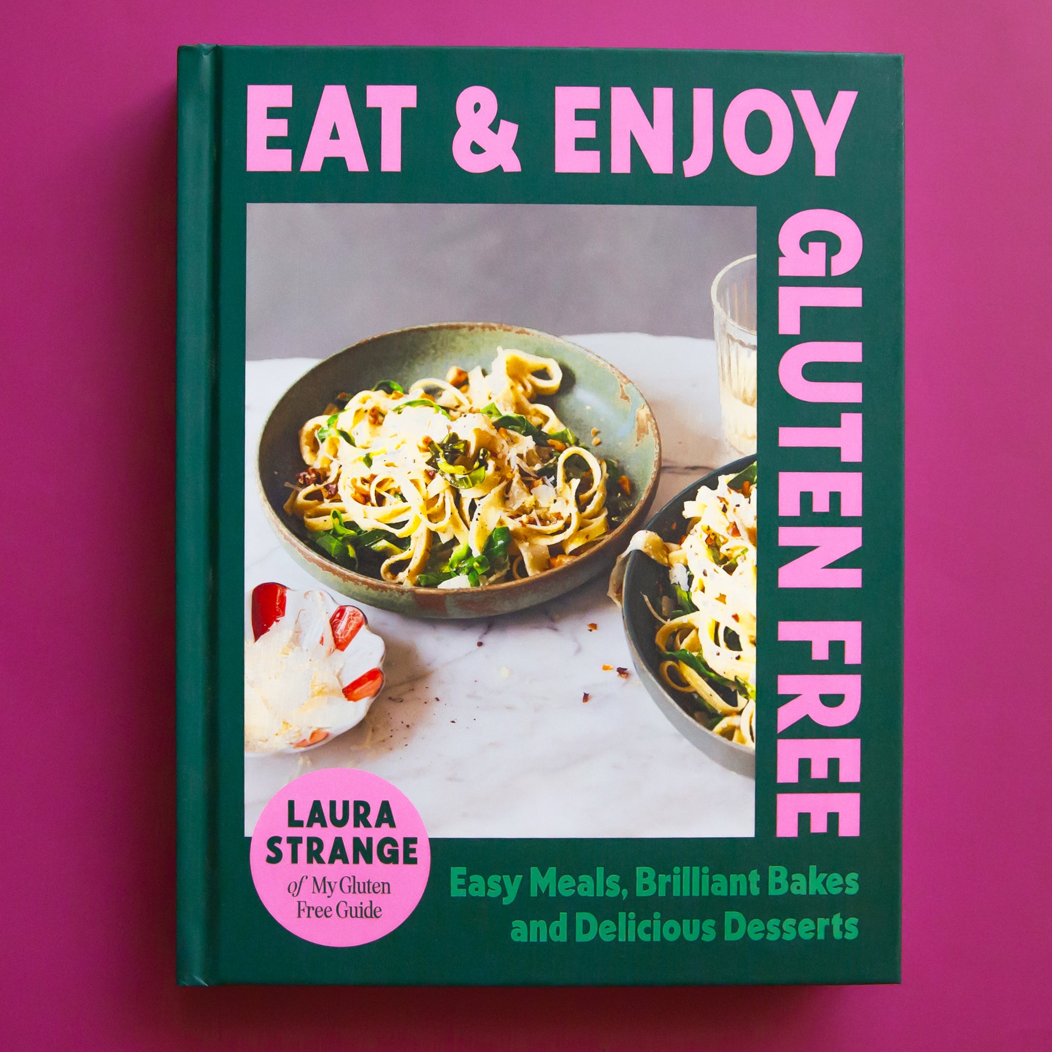 On a purple background is a green and purple book cover with a photo of food on the front and the title around the edges that reads, &quot;Eat &amp; Enjoy Gluten Free&quot;. 