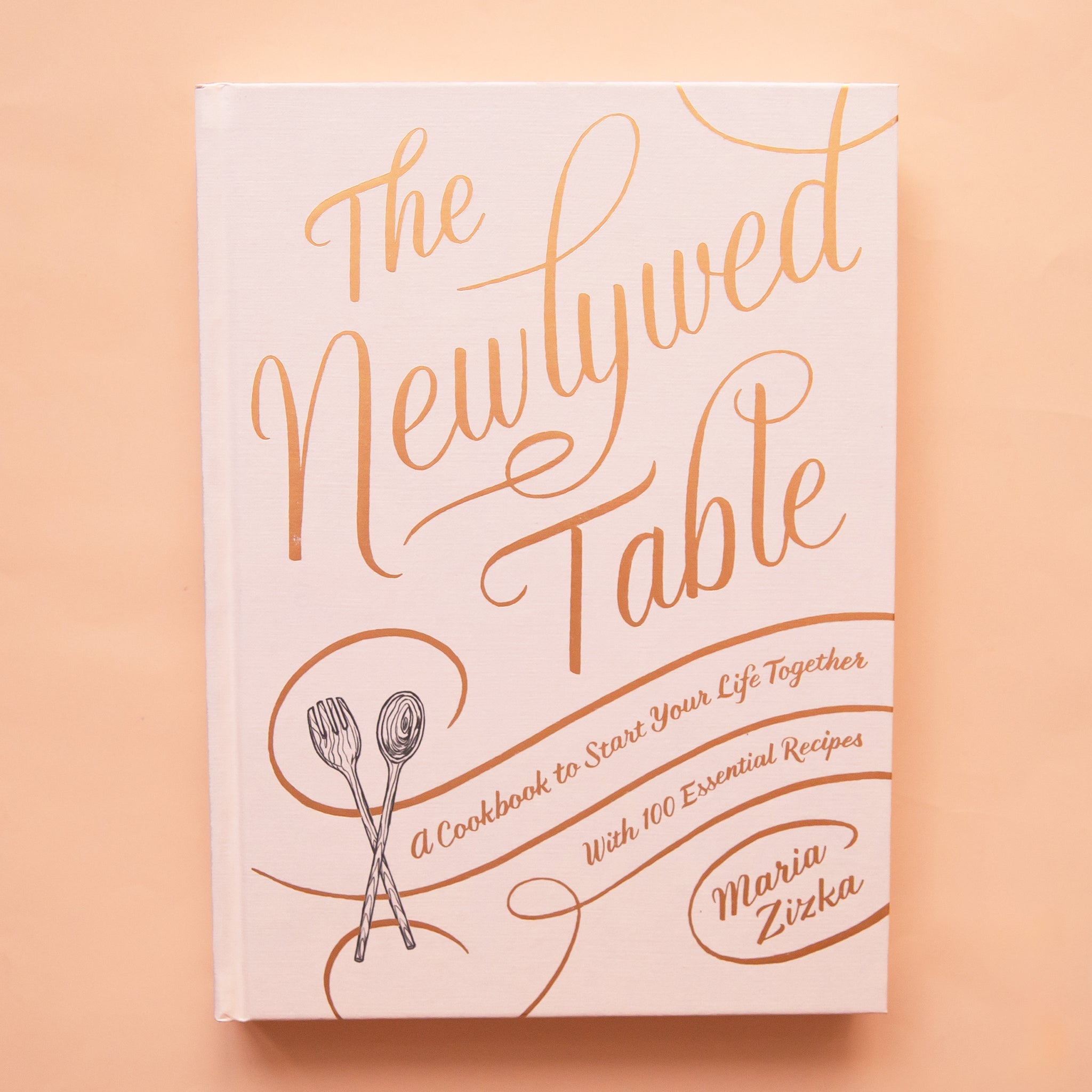White hardcover book titled &#39;The newlywed table&#39; in gold foil cursive lettering. Below reads &#39;a cookbook to start your life together with 100 essential recipes&#39;. A black and white wooden spork and spoon sits in the bottom left corner.