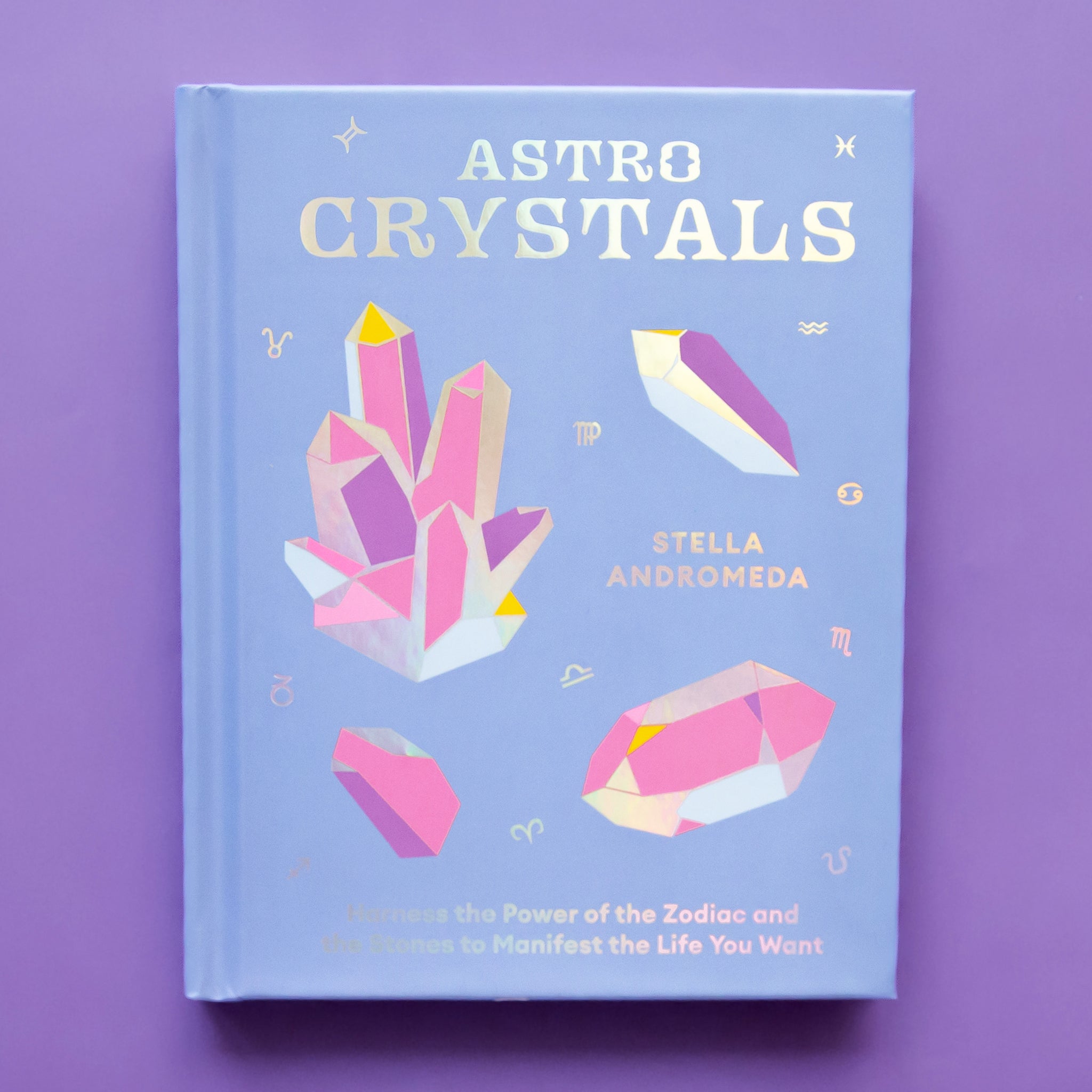On a purple background is a periwinkle colored book cover with graphics of crystals and clusters and the title that reads, "Astro Crystals". 