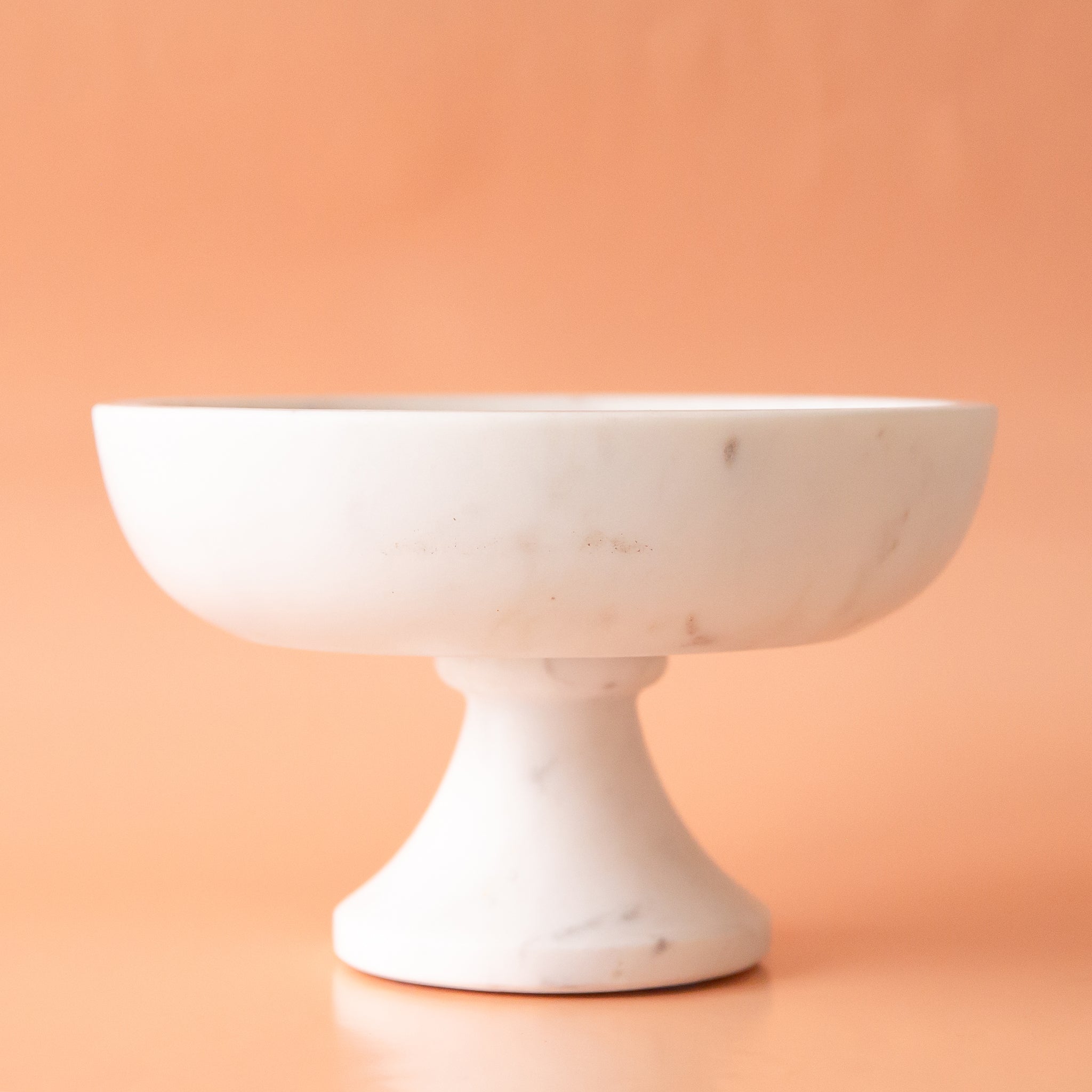 On a peachy background is a white marble pedestal bowl. 