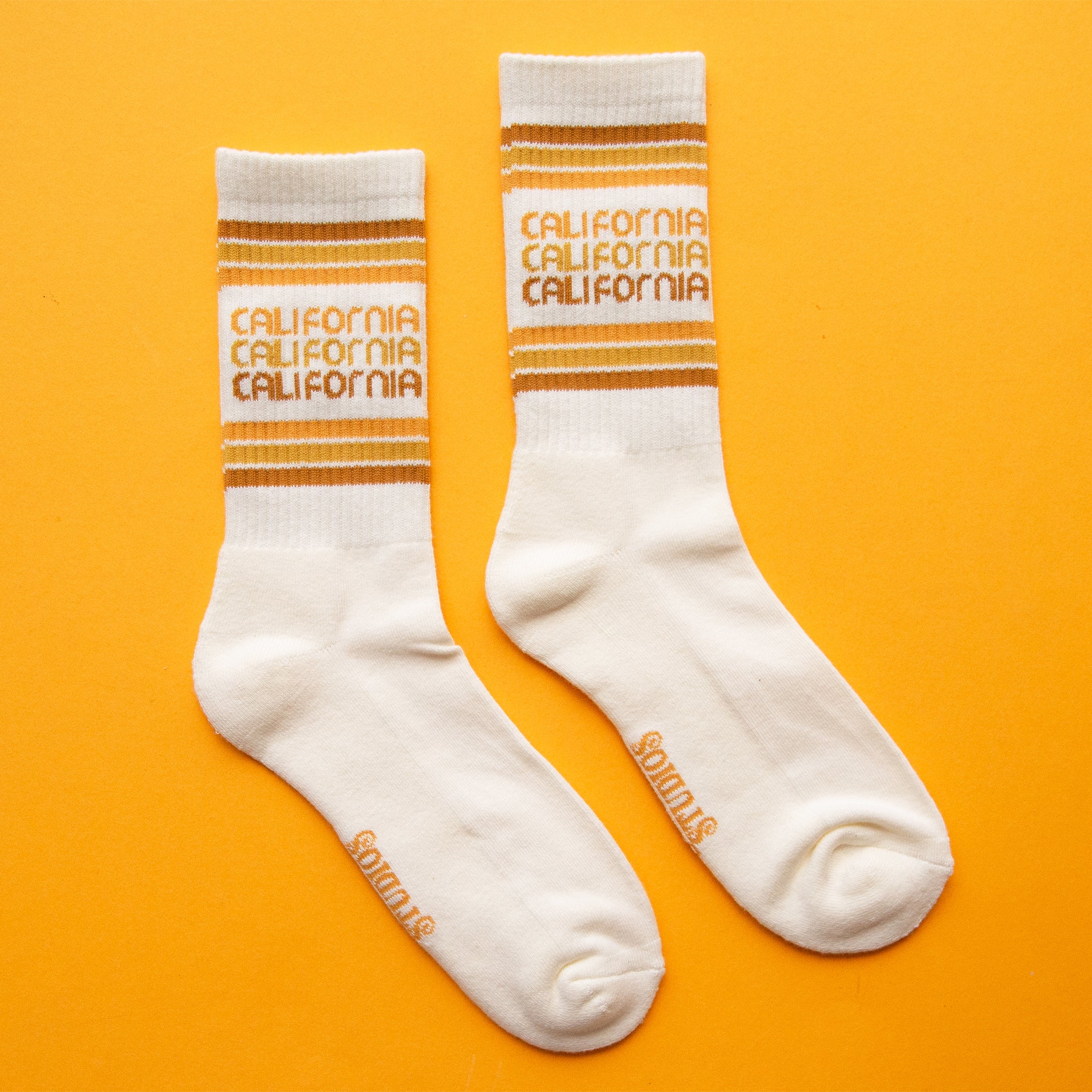 On a yellow background is a white socks with rust, yellow and orange stripes and "california" printed three times on the ankle.