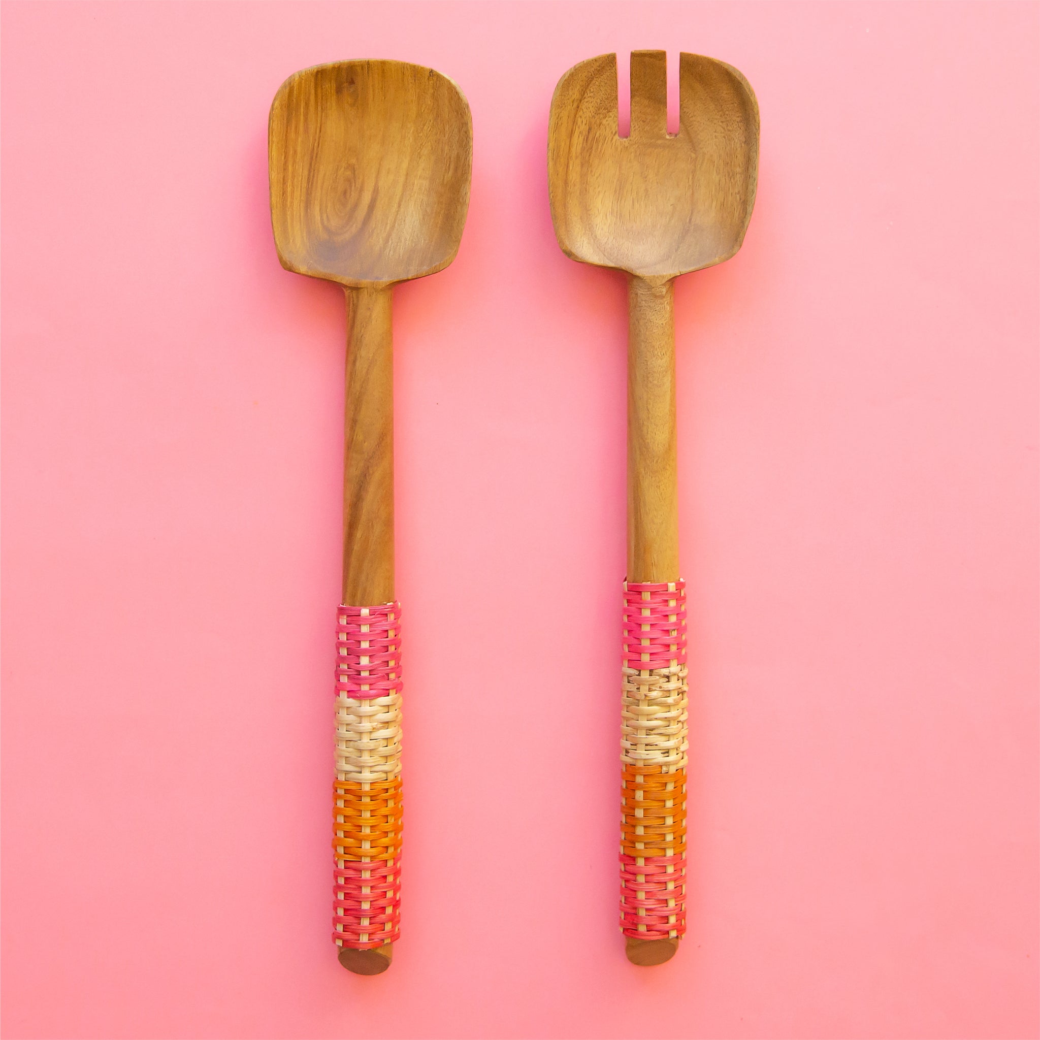 On a pink background is a pair of wooden salad servers with long handles wrapped in orange, pink and neutral colored rattan. 