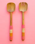 On a pink background is a pair of wooden salad servers with long handles wrapped in orange, pink and neutral colored rattan.