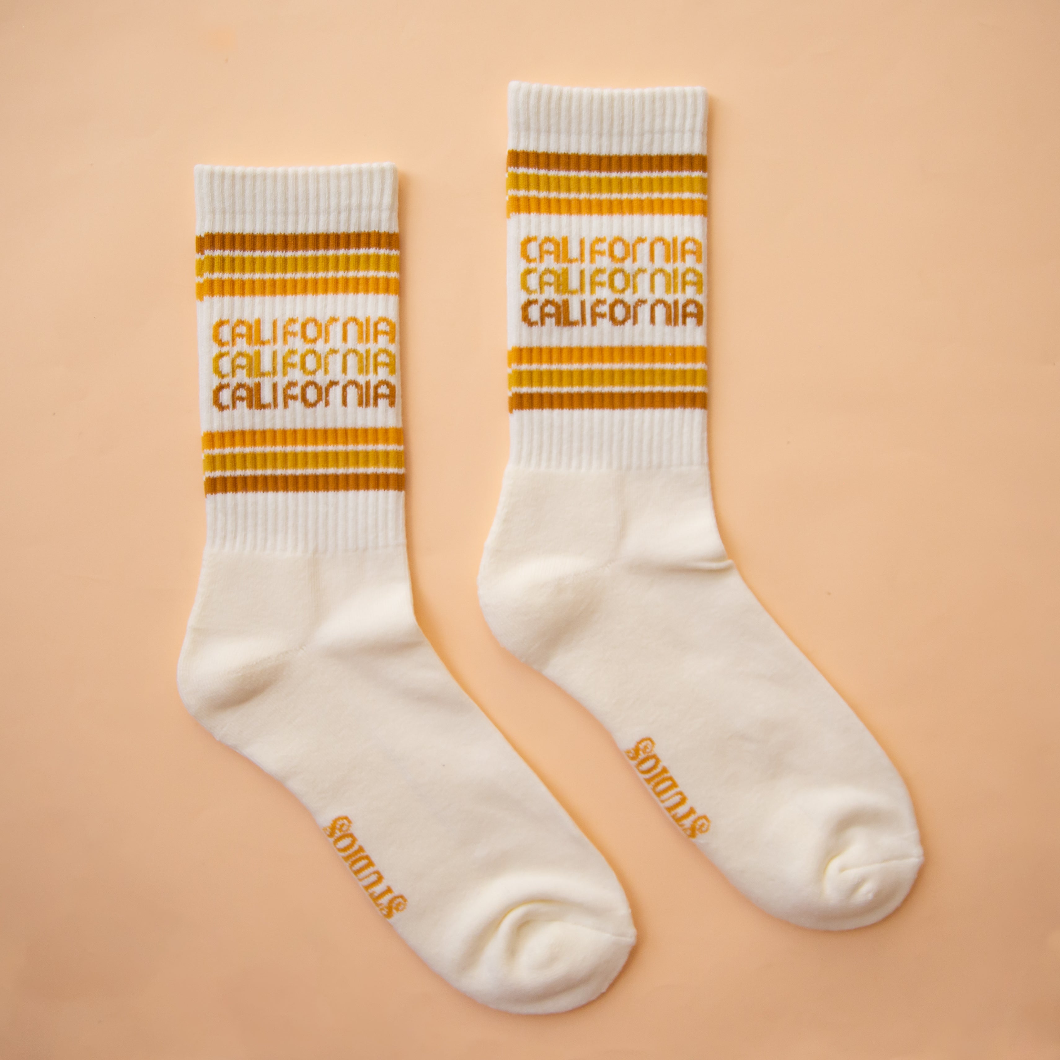 On a peach background is a white socks with rust, yellow and orange stripes and "california" printed three times on the ankle. 