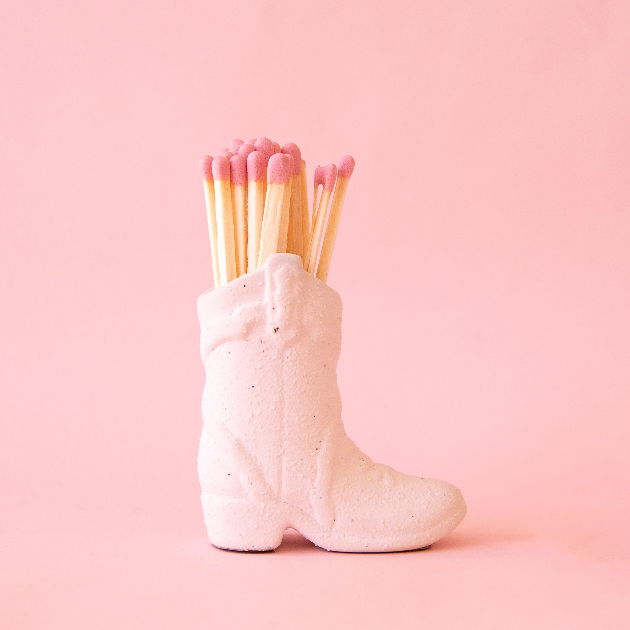 On a pink background is a white cowgirl boot shaped match holder with pink matches inside