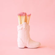 On a pink background is a white cowgirl boot shaped match holder with pink matches inside