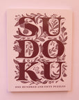 Pastel pink paperback puzzle book titled 'Sudoku' in large magenta capital lettering. The letters are accented with delightful floral detailing. Below the large lettering reads 'One hundred and fifty puzzles' towards the bottom of the cover.
