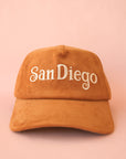 On a pink background is a toffee colored baseball cap that has a corduroy texture and says, "San Diego" in white lettering.