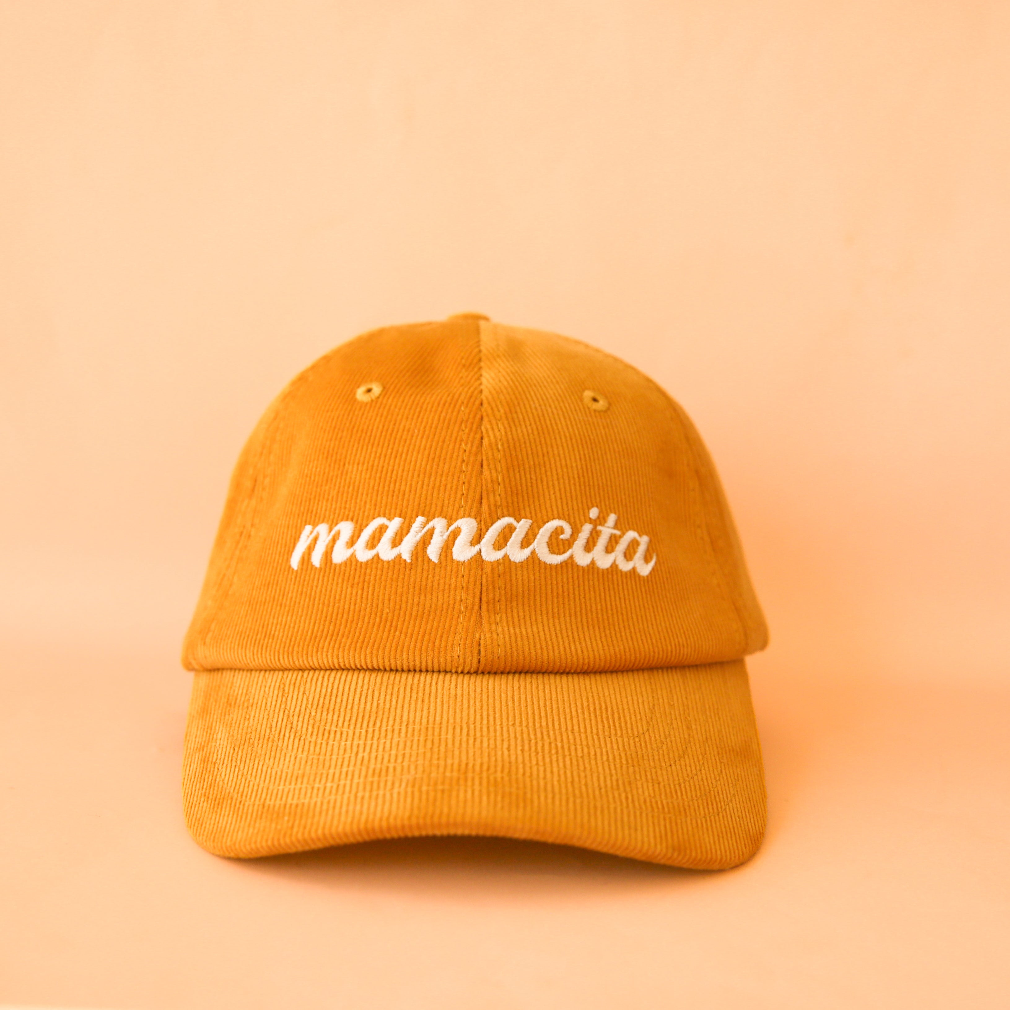 On an orange background is a corduroy baseball hat with white embroidery that reads, &quot;mamacita&quot;. 