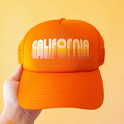On a yellow background is an orange trucker hat with yellow text across the front that reads, "California".