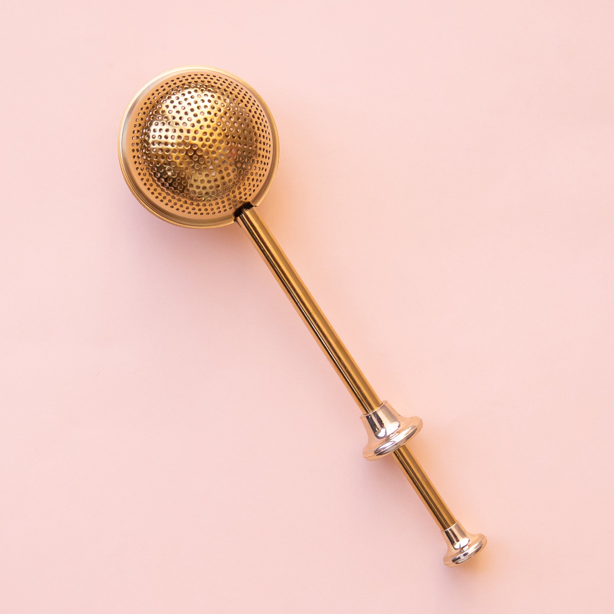 On a pink background is a gold stainless steel tea infusing egg. 