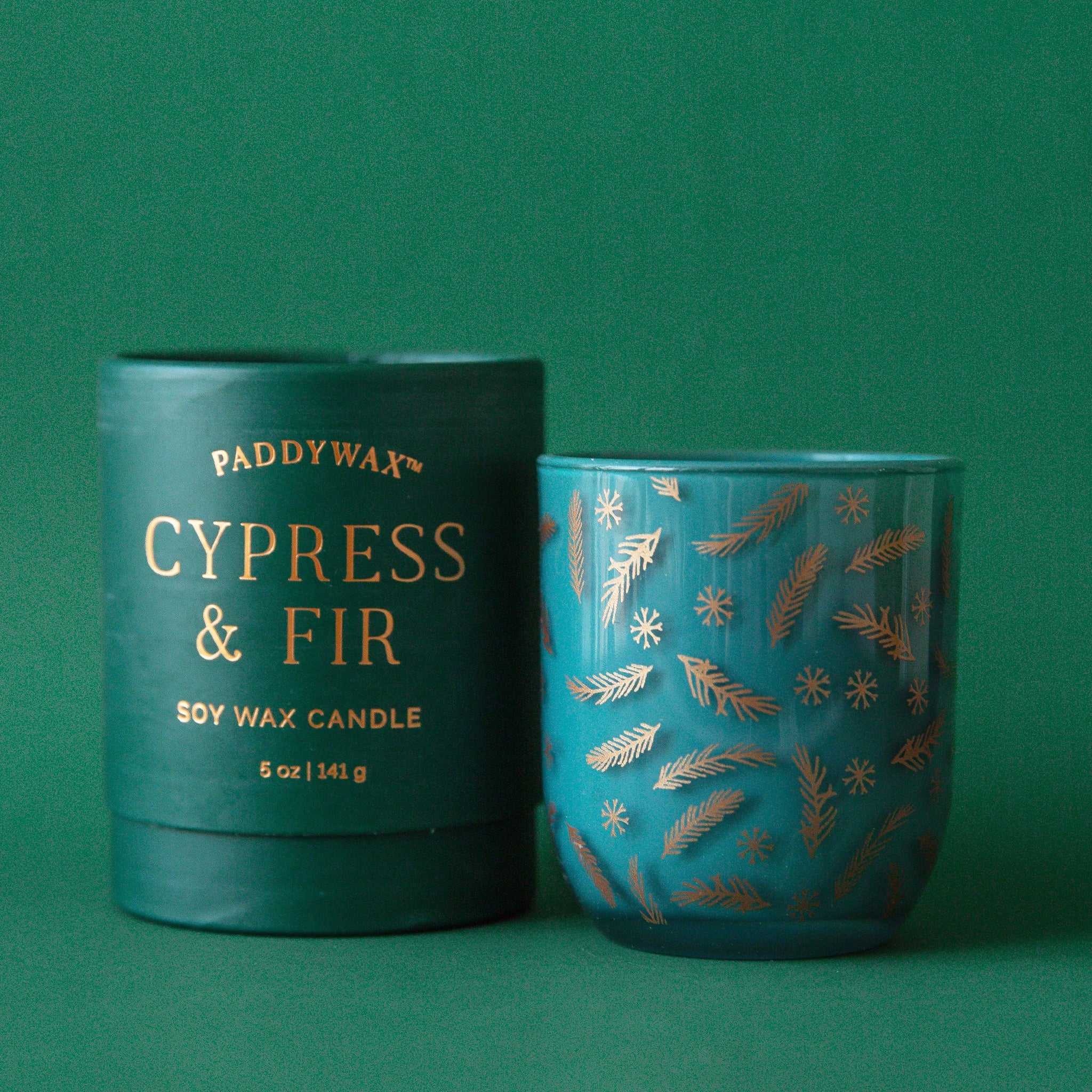 On a dark green background is a glass candle jar in a dark green shade with gold pine and snowflake designs alongside a cardboard tube container with gold foiled text that reads, "Paddywax Cypress & Fir Soy Wax Candle".