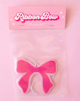 A hot pink bow with light pink trim air freshener is shown against a light pink to peach gradient background.