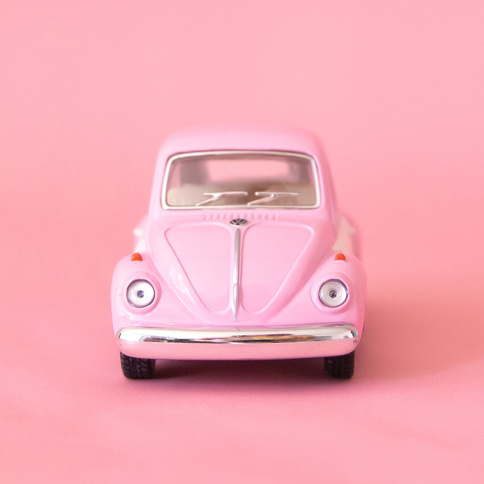On a pink background is a pink and white two toned toy in the shape of a volkswagen beetle.
