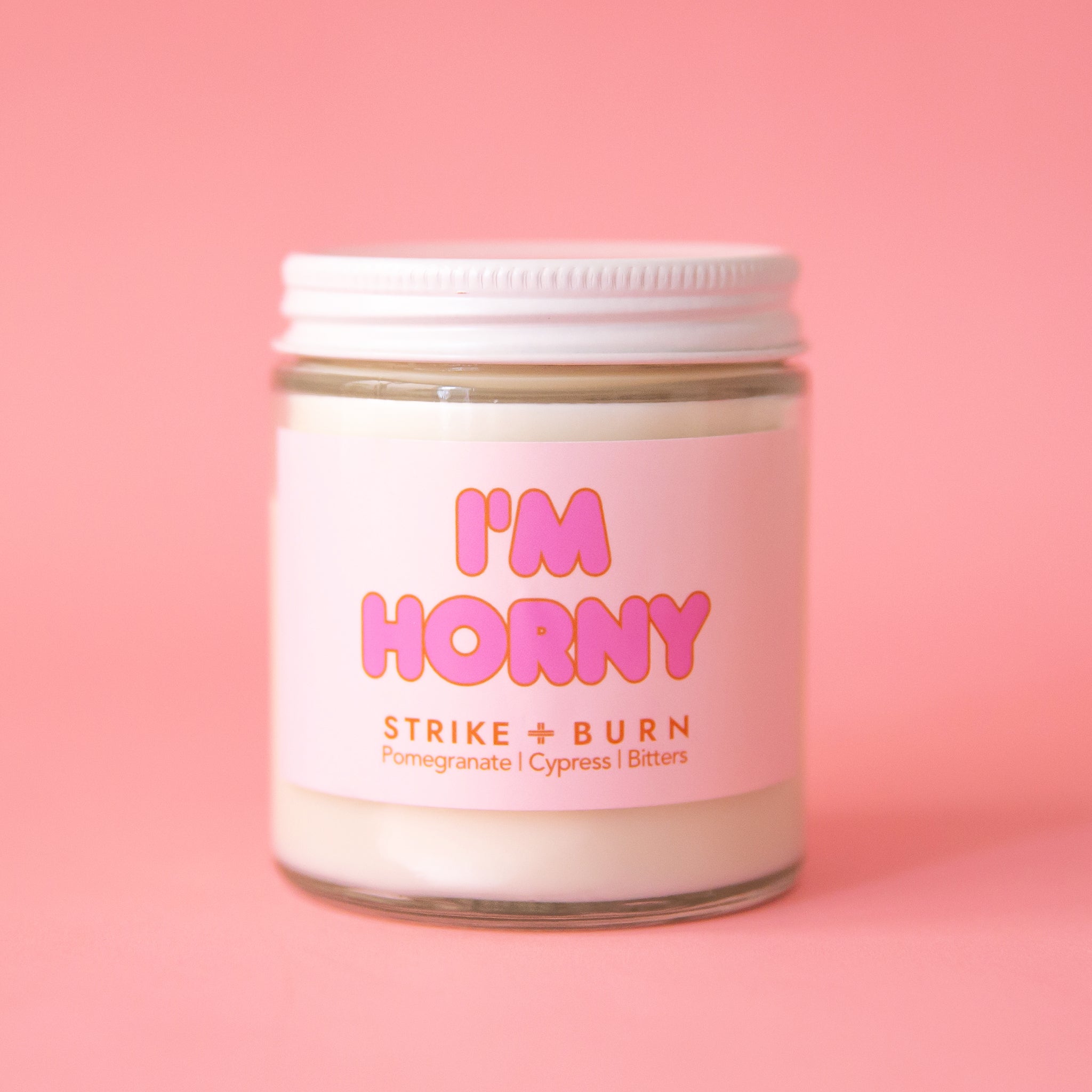 On a pink background is a clear glass jar candle with a lid and label that read, "I'm Horny". 