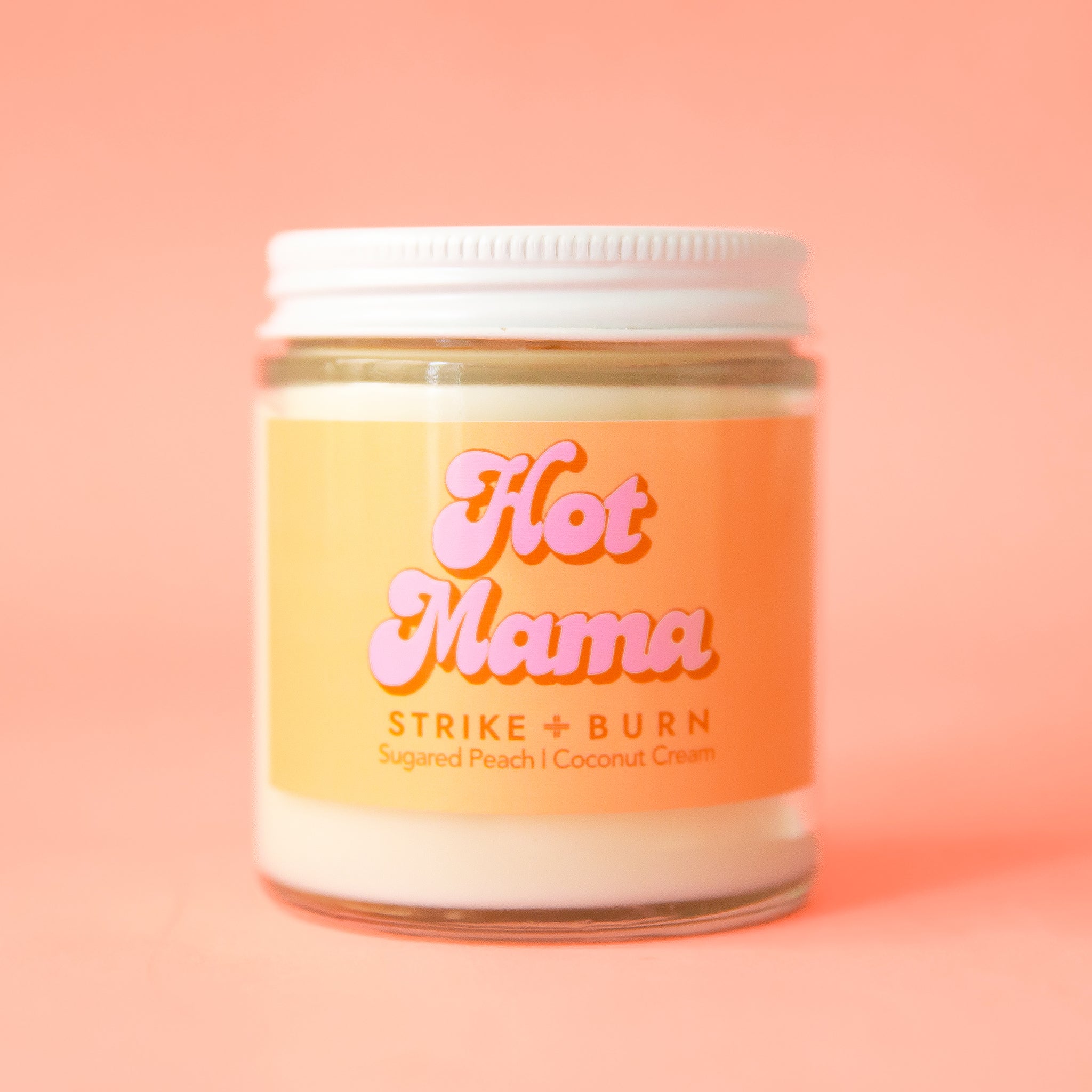 A clear glass jar candle with an orange and pink label that reads, "Hot Mama".