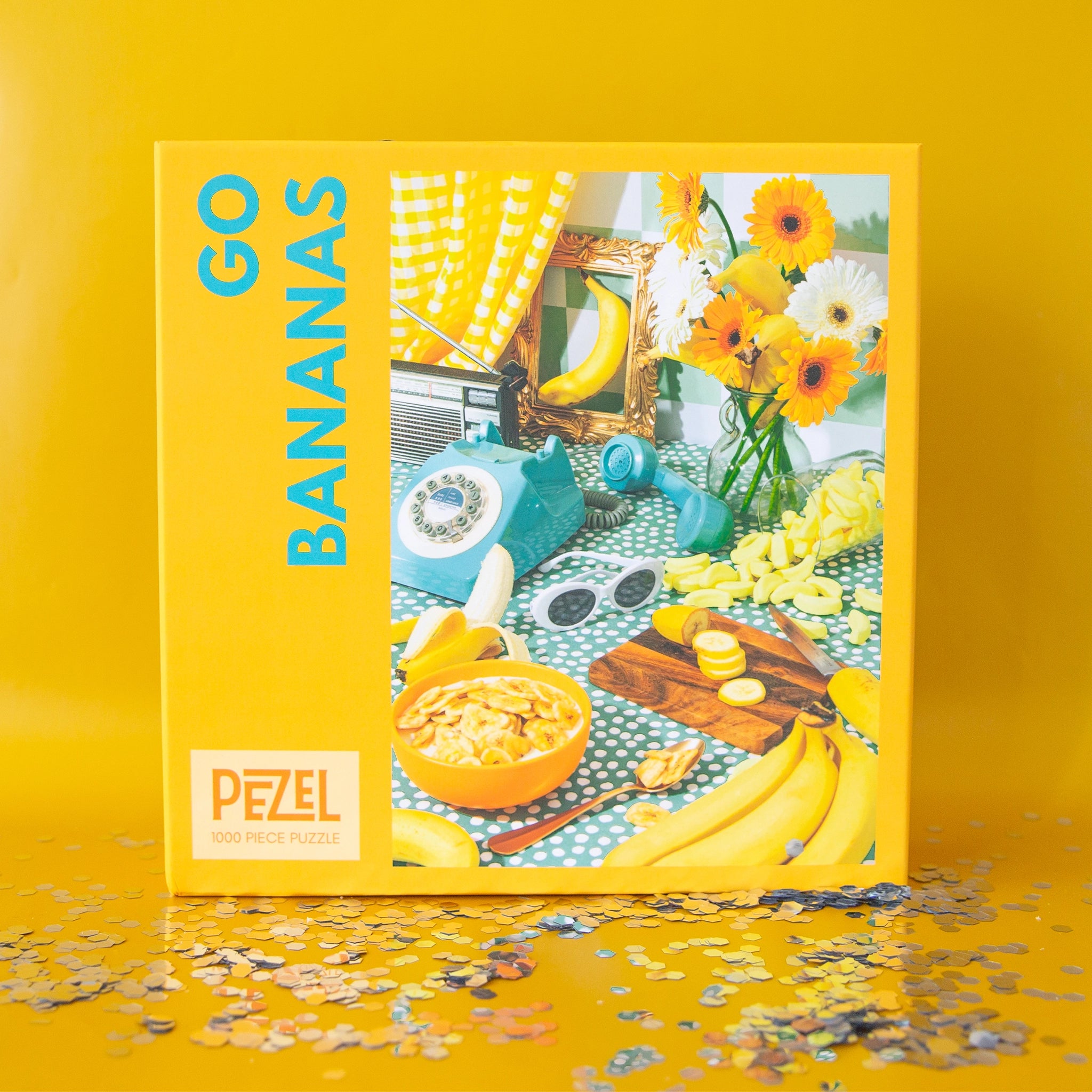 On a yellow background is a red and blue puzzle box ghat reads, &quot;Go Bananas&quot; with a photo on the side of what the puzzle looks like finished which is a retro table scape with bananas a vintage touch dial phone yellow flowers and white oval sunglasses. 