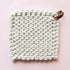 On a light pink background is a square crocheted pot holder in a cream shade and a brown leather loop on the corner for hanging. 