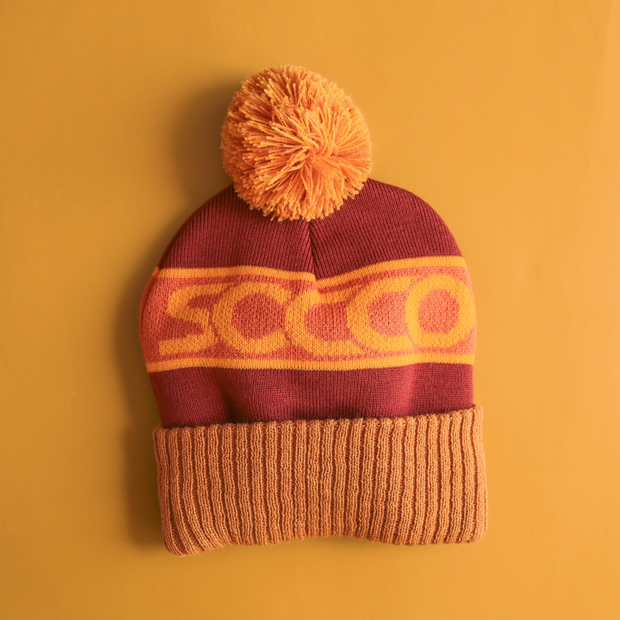 On a yellow background is a yellow and rust colored beanie with a pom pom on top and "SOCCO" written across the center. 