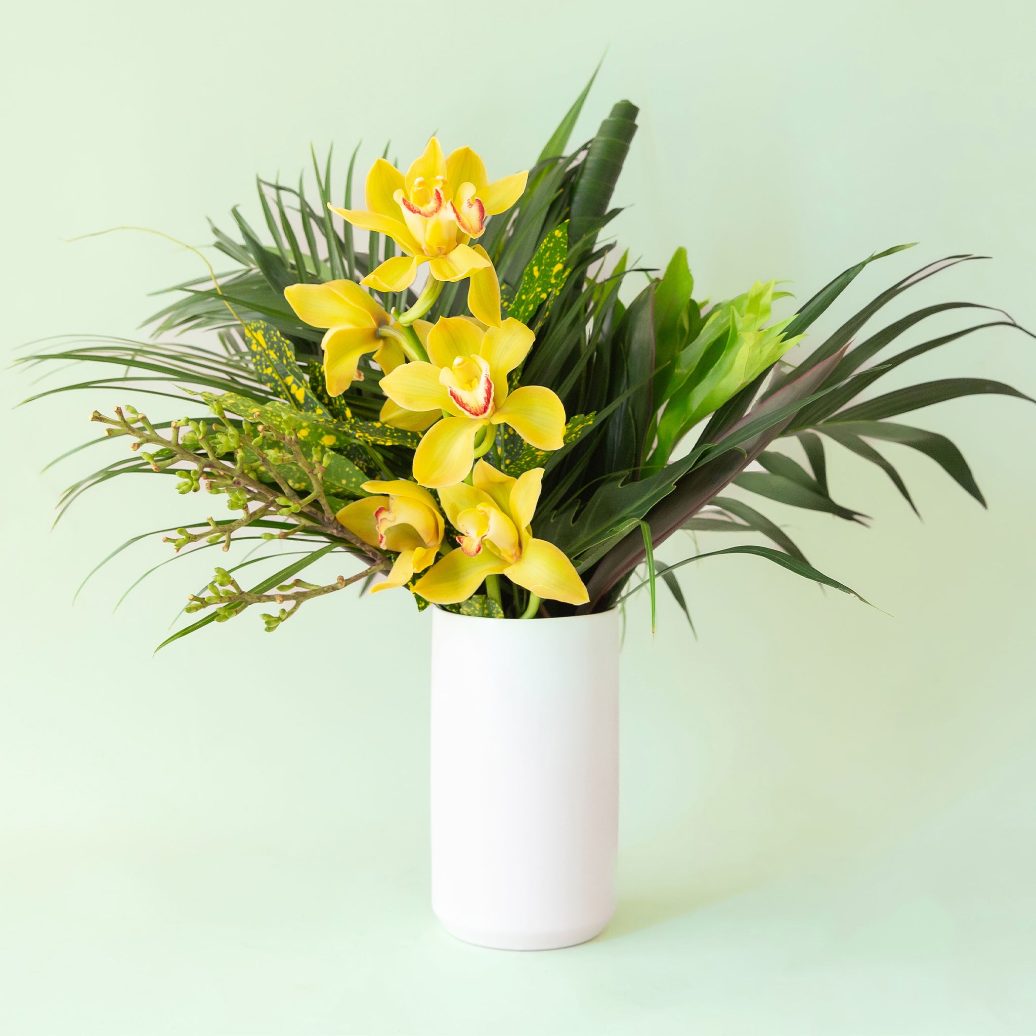 Narrow white cylinder vase filled with palms and tropical flowers not included with purchase. The vase lays against a light green background.