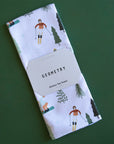 On a green background is a folded white waffle knit kitchen towel with a ski and snowboard pattern.