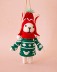 On a peachy background is a white felt dog ornament with a green sweater and red hat on.