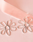 A pair of silver floral earrings lay on a pink background
