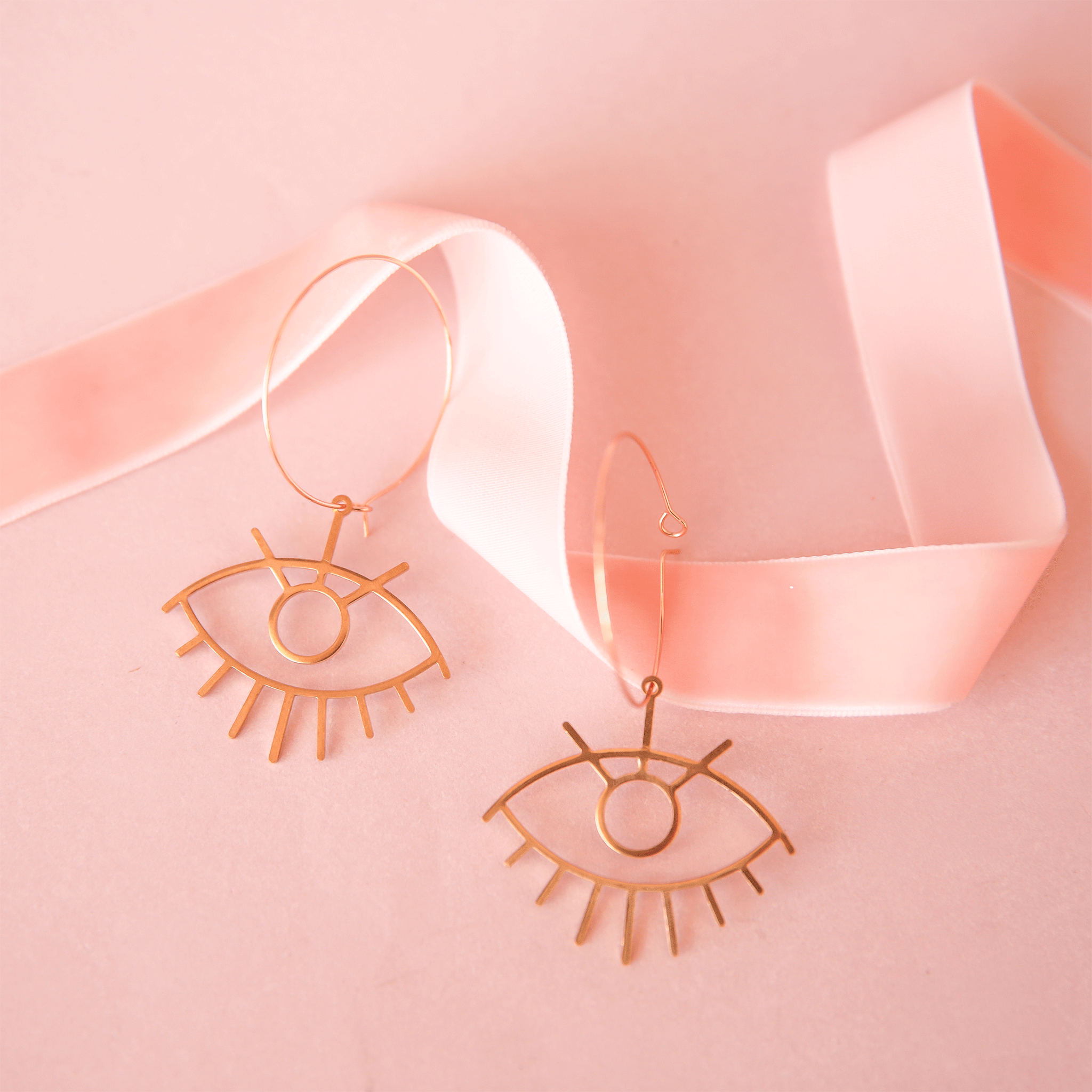 On a pink background is a pair of thin gold hoops earrings and an eye shaped charm hanging from the bottom of the hoops. 