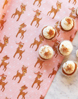 A pink kitchen towel with a brown retro deer pattern and staged next to white frosted cupcakes.